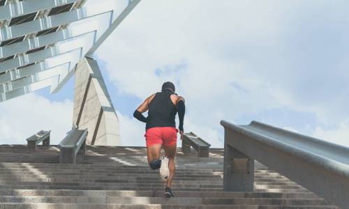 Muscular hispanic Young male dark-skinned athlete running up a flight of stairs with speed, sporty young man in black t-shirt training or working out outdoors while jogging up the steps. Rear view.